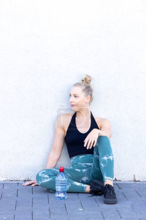 A happy and sporty young blonde millennial woman sits on the floor with a bottle of water after exercising, radiating wellness and a commitment to a healthy lifestyle while looking at the camera