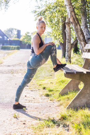 A young adult female in athletic wear engages in a stretching exercise by placing her foot on a wooden park bench. She exhibits focus and determination amid a sunlit, natural setting with birch trees