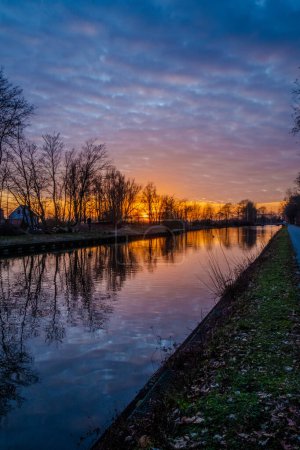 This image presents a striking sunset along a calm river, where the sky is adorned with an array of colors ranging from deep blue to fiery orange. The silhouettes of bare trees line the riverbank
