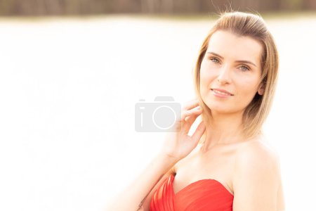 A portrait of a poised woman dressed in an elegant red strapless dress, bathed in the warm, soft glow of sunlight. Her confident gaze and gentle touch on her shoulder express a sense of ease and