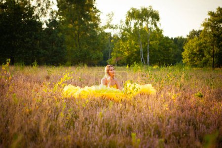 Capturada a la hora dorada, esta imagen muestra a una joven sentada en un campo, envuelta por el cálido abrazo del atardecer. Su vestido amarillo se extiende a su alrededor como un charco de luz solar, en contraste con