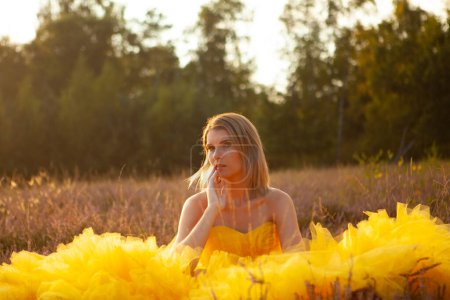 Inmitten eines Feldes bei Sonnenuntergang sitzt eine Frau nachdenklich, ihr voluminöses gelbes Kleid weht um sie herum. Das goldene Stundenlicht erhellt sanft ihren nachdenklichen Ausdruck, und der natürliche Hintergrund verblasst.