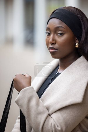 En este primer plano, una joven mujer negra emana elegancia urbana, vestida con un elegante abrigo crema sobre un traje negro clásico. Su diadema y pendientes de oro añaden un toque de sofisticación, mientras que su mirada
