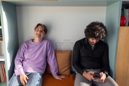 Esta foto captura el contraste de las interacciones sociales modernas entre amigos. Por un lado, una persona en un suéter de lavanda se ríe de todo corazón, mientras que por el otro, su amigo con el pelo rizado y un negro