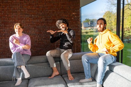 In einem sonnendurchfluteten Raum mit großen Fenstern und einer Ziegelwand sitzen drei barfüßige Menschen auf einem grauen Sofa. Sie zeigen animierte Mimik und Gestik, die eine lebendige Interaktion suggerieren. Die Person