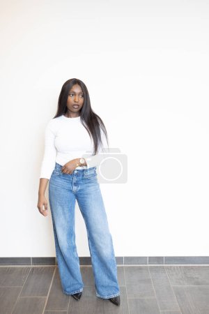 A young Black woman showcases a timeless, trendy look in a fitted white top paired with classic blue jeans. Her hand rests on her hip, a gold watch gleaming as an accent. She stands against a stark