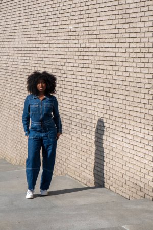 A woman stands casually against a pale brick wall, her face accidentally obscured by a watermark in the image. Shes dressed in a stylish denim jumpsuit paired with white sneakers, embodying a relaxed