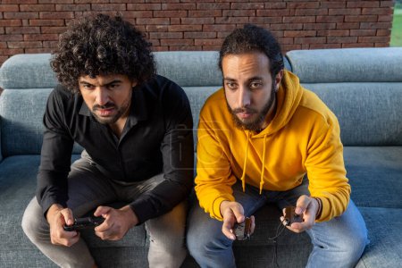 Photo for Two engaged adult males are seated on a grey couch, holding game controllers with intense concentration. They are indoors, with a red brick wall in the background, implying a casual, home environment - Royalty Free Image