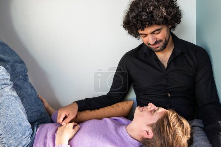 Esta cálida fotografía captura un momento íntimo entre una pareja en la comodidad de su hogar. Un hombre con el pelo rizado y una camisa negra sonríe cariñosamente mientras mira a su compañero, que yace relajado