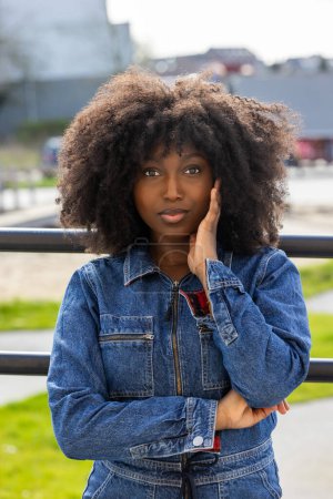 This image portrays a young Black woman leaning against a railing on a city bridge, lost in thought. Her large afro and denim jacket symbolize a blend of urban style and introspection. The unfocused
