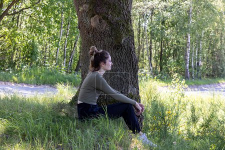 This image eloquently captures a young woman in a contemplative state, sitting against an old trees sturdy trunk. Her profile is set against a backdrop of dense birch trees and lush undergrowth, with