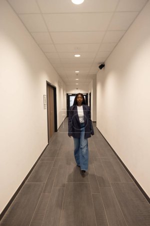 An African American woman with a medium build is featured walking toward the camera in a well-lit office corridor. She has a relaxed posture, conveying confidence. Her style is casual-professional