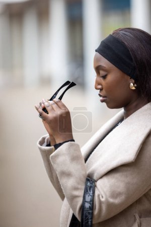 An African American woman is depicted examining a pair of sunglasses outdoors. Shes dressed in a light beige coat with a black belt and headband, exuding a sense of casual urban fashion. The shallow