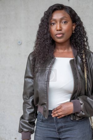 A poised African American woman stands against a concrete wall, exuding confidence. Her curly black hair frames her face, complemented by subtle makeup. Shes clad in a modern ensemble featuring a
