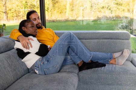 Una pareja multirracial disfruta de un momento tranquilo e íntimo juntos, descansando cómodamente en un sofá gris. Están en un abrazo pacífico, vestidos casualmente con denim y suéteres suaves, con un brillante, natural