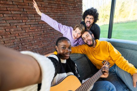 Capturant un moment de pure joie, cette image montre un groupe d'amis divers prenant un selfie. L'un joue de la guitare, tandis que les autres montrent des expressions ludiques. Ils sont assis contre un mur de briques à l'intérieur