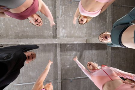This image captures a unique low-angle view from beneath a circle of women dressed in athletic attire within a spacious concrete setting. Their expressions are lively and joyful, suggesting a moment