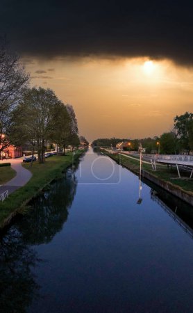 As twilight descends, a canal stretches into the distance, flanked by trees and the warm glow of street lights. The setting sun pierces through the heavy clouds, casting a dramatic light over the