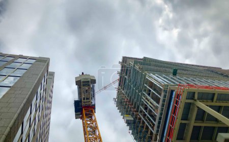 La imagen captura la expansión ascendente de un paisaje urbano, yuxtaponiendo los elegantes exteriores de los rascacielos terminados con el marco en bruto de un edificio en construcción. Una grúa imponente