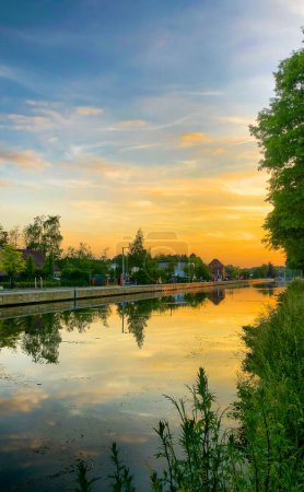 This image features a tranquil canal scene at sunset. The waterway mirrors the sky, with hues of orange and blue, and a gradient sky. Flora frames the waters edge. In the background, a small village