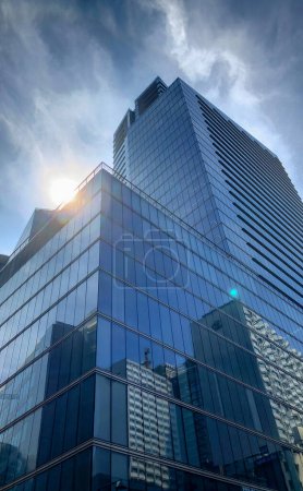 Foto de La imagen captura la imponente arquitectura de un rascacielos moderno. Su fachada de vidrio reflectante brilla con los rayos solares, creando un efecto de destello de lente. El cielo azul, salpicado de nubes, contrasta - Imagen libre de derechos