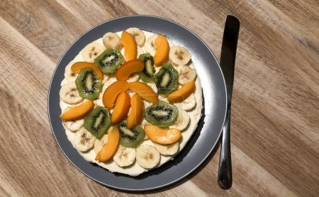 Displayed is a colorful fruit arrangement on a plate, featuring neatly sliced bananas, vibrant green kiwi, and soft orange apricots, all resting on a creamy bed of yogurt. The wooden tabletop adds