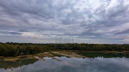 The photograph presents a vast landscape where a still lake reflects the complex patterns of a brooding sky, heavy with clouds. The surrounding forest, a lush canvas of greens, anchors the scene