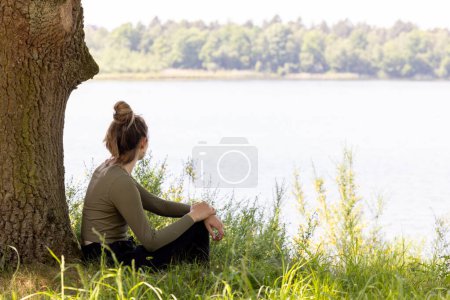 Eine junge Frau sitzt kontemplativ an einem ruhigen See, umgeben von der Bequemlichkeit des Grüns der Natur und einem großen, strukturierten Baum. Ihre entspannte Haltung und das ruhige Wasser im Hintergrund schaffen eine