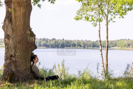 An image of quiet repose, showing a woman seated under the sturdy trunk of a tree. The lake in the background, framed by verdant foliage, accentuates the feeling of solitude and the connection with