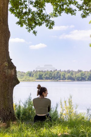 In dieser ruhigen Umgebung sitzt eine Frau nachdenklich unter dem Baldachin eines Baumes mit Blick auf einen ruhigen See. Die Komposition balanciert die Größe der Natur mit einem persönlichen, introspektiven menschlichen Element aus