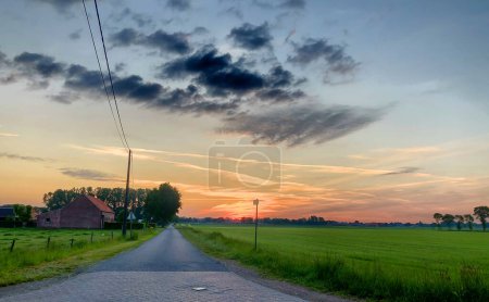This image captures the breathtaking beauty of a sunrise over a rural landscape, featuring a long road leading past a traditional farmhouse surrounded by lush green fields. The sky, painted with
