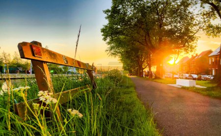 This image captures the essence of a tranquil suburban evening, showcasing a path lined with lush grass and wildflowers, with a rustic wooden barrier in the foreground. The setting sun casts a warm