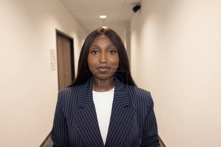 This professional portrait features a young Black woman confidently standing in a corporate hallway. She is dressed in a sophisticated striped suit with a crisp white blouse, embodying both style and