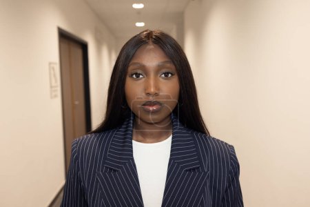 An African American woman stands confidently in a brightly lit office corridor, dressed in a stylish striped business suit. She gazes directly at the camera with a neutral expression, her long