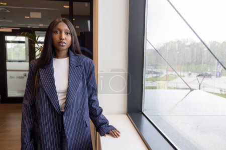 Esta imagen captura a una joven afroamericana en un entorno profesional, vestida con un traje de negocios a rayas, de pie junto a una gran ventana con vistas a un día lluvioso afuera. Su comportamiento calmado