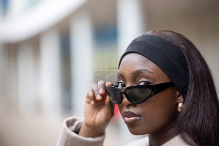 A close-up portrait of a stylish African American woman adjusting her oversized sunglasses outdoors. Her attire is casual yet chic, complemented by subtle makeup and a black headband. The blurred