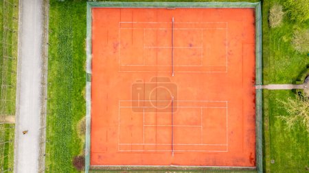 This aerial image features an unoccupied red clay tennis court, distinct with its bright playing surface and crisp white lines. Enclosed by lush green grass and a narrow path, the court stands out in