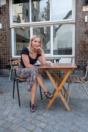 Captured in this image is a serene, blonde woman sitting alone at a wooden cafe table on a cobblestone sidewalk. She leans gently on her elbow, her chin resting on her hand, portraying a pensive