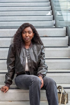 A young Black woman sits casually on concrete steps in an urban environment. She wears a stylish black leather jacket over a white tank top, paired with faded jeans, and a tan and black woven tote bag