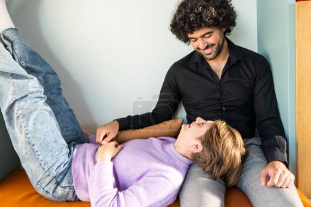 Capturing a serene moment, this image features two friends in a comforting and casual environment. An androgynous person wearing a lavender sweater lies down with legs up against the wall, while a