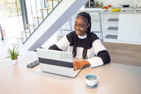 A cheerful young Black woman works on her laptop in a bright, stylish kitchen. The setting includes a modern staircase and a neat, contemporary kitchen background. She wears a cozy black and white