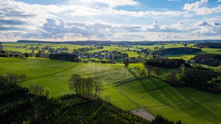 This aerial photograph captures the vast and verdant landscape of the Hautes Fagnes region, characterized by its rolling hills and vibrant green fields. The image showcases the areas natural beauty