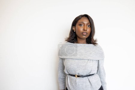 Dieses Bild zeigt eine junge schwarze Frau, die vor einem schlichten weißen Hintergrund steht, in einem schicken grauen Pullover gekleidet und an der Taille angeschnallt. Ihr Gesichtsausdruck ist ruhig und nachdenklich und vermittelt ein Gefühl von