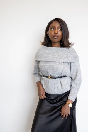 This portrait captures a young Black woman posed against a plain white background, embodying sophistication and confidence. She wears a light grey, cropped sweater and a sleek black leather skirt
