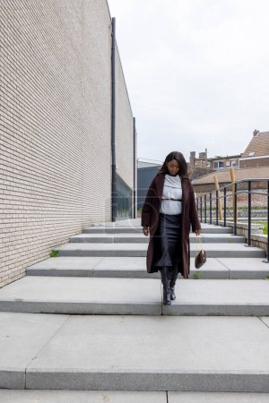 This image captures a young Black woman descending outdoor steps in an urban environment, exuding confidence with every step. She is dressed in a stylish winter ensemble, including a long brown coat