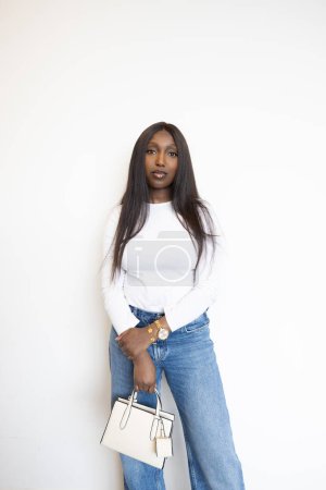 This portrait features a young Black woman standing against a plain white background, exuding elegance and confidence. She is dressed in a classic white long-sleeve top and casual blue jeans