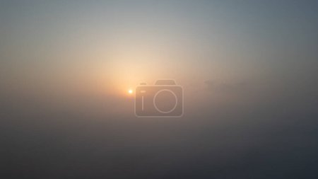 This image captures a serene sunrise, with the sun emerging softly through a dense morning fog. The muted palette of the scene conveys a tranquil and peaceful atmosphere, as the suns gentle glow