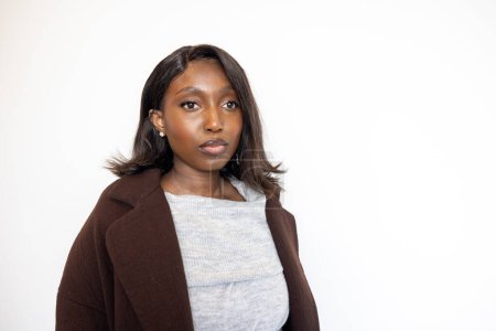 This portrait features a confident young Black woman dressed in stylish winter attire. She wears a gray turtleneck sweater and a dark brown coat, with her dark hair flowing around her shoulders. The