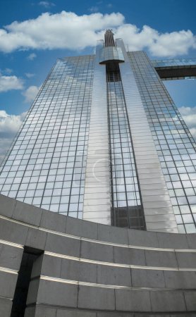 The impressive facade of a modern skyscraper reaches skyward, its surface reflecting the bright day. Fluffy white clouds scatter across the blue sky, creating a dynamic contrast with the sleek