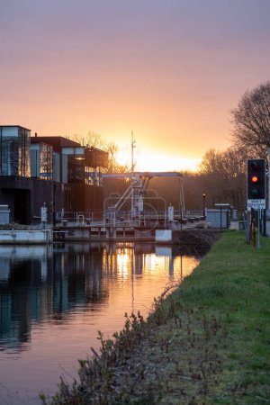 As the sun sets, its radiant glow bathes an industrial waterway and adjacent modern buildings in warm light. The sunset reflects on the calm water, creating a harmonious blend of nature and industry